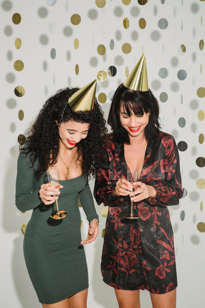 Positive female friends in dresses wearing party hats looking down while standing with sparkling alcohol drink in wineglasses on white background with paper garland